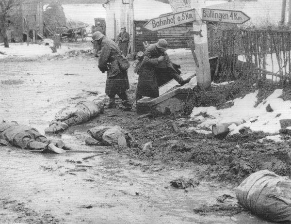 Jeńcy amerykańscy pomordowani w Honsfeld przez Kampfgruppe Peiper. Niemiecki żołnierz przymierza „zdobyczne” buty (fot. U.S. Army, ze zbiorów National Archives and Records Administration, domena publiczna).